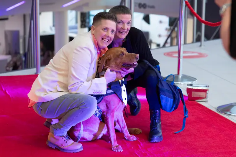 Two guests crouching on the red carpet beside a dog in a bow tie.