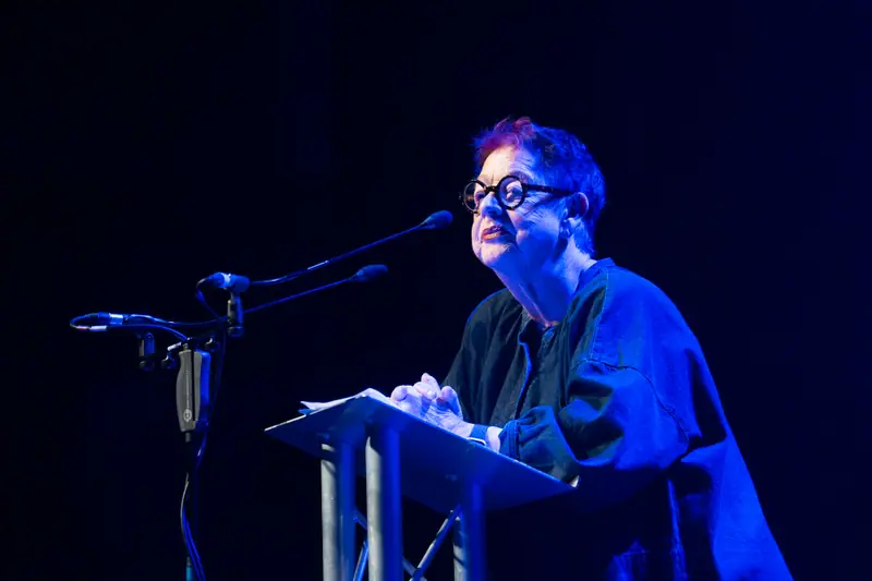 Jo Brand reading from the lectern under a blue stage wash.