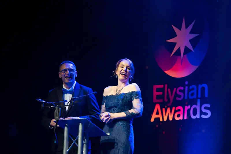 Two hosts laughing on stage beside the Elysian Awards wordmark.